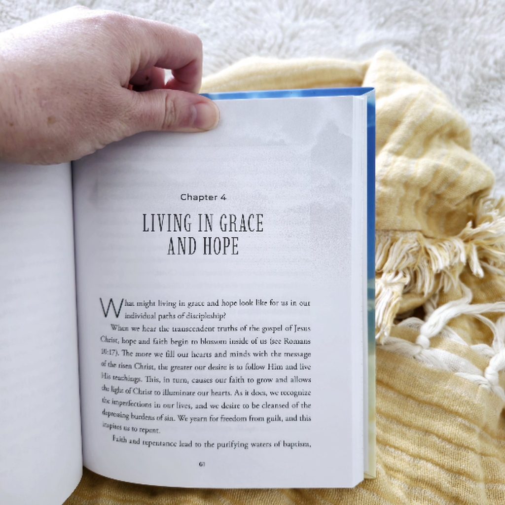 A close-up of an open book showing Chapter 4, titled Living in Grace and Hope. A person's left hand holds the top left corner of the page. The text on the page discusses discipleship, faith, repentance, and the message of Jesus Christ. The page has transparent clous decorating the top. The book rests on a soft, mustard-yellow throw blanket with white fringe, which is draped over a plush light gray surface. The cozy setting suggests a peaceful reading moment.