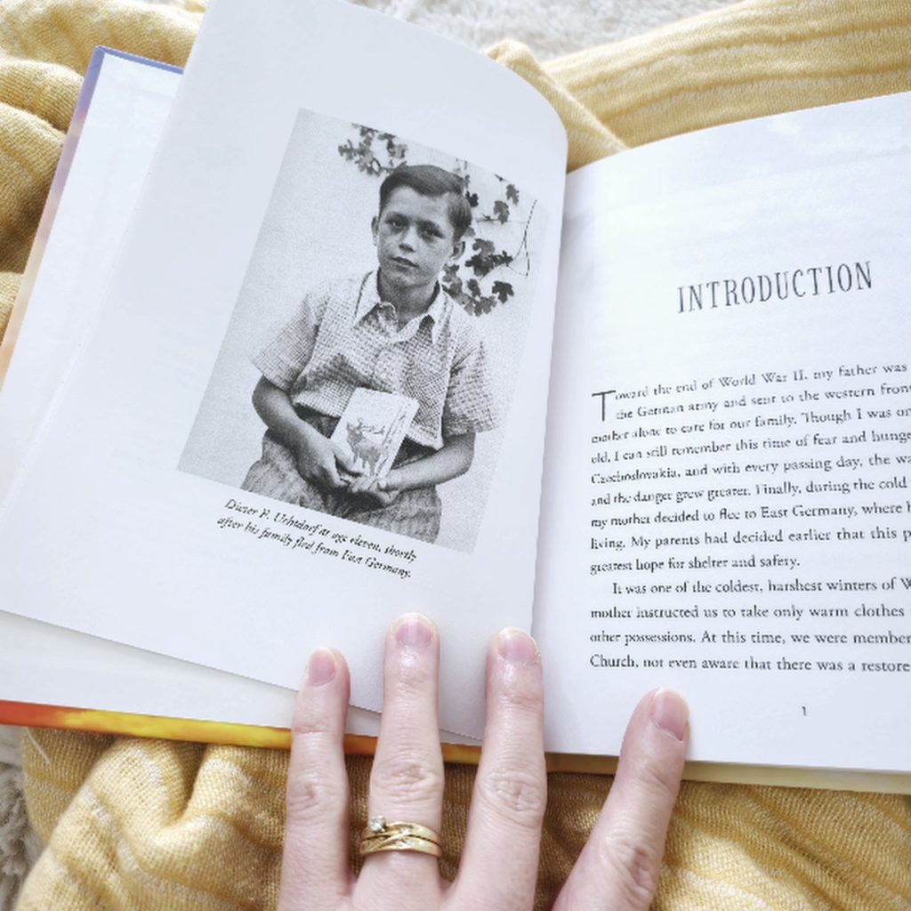 An open book displays the introduction page and a black-and-white photograph of a young Dieter F. Uchtdorf at age eleven, holding a book, with a caption stating that this was after his family fled from East Germany. The introduction text discusses World War II and his family's escape to East Germany. A person's left hand, wearing a gold wedding ring, gently holds the bottom of the page. The book rests on a mustard-yellow throw blanket with white fringe, adding a cozy and warm atmosphere to the scene.
