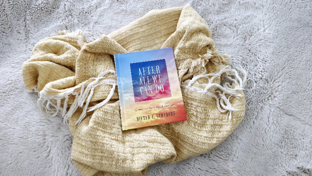 A book titled After All We Can Do by Dieter F. Uchtdorf rests on a soft, light cream plush blanket. The book cover features a gradient sky with hues of blue, pink, and orange. The book is partially draped over a cozy, textured mustard-yellow throw blanket with white fringe, which is casually arranged. The scene gives a warm, inviting atmosphere, suggesting a peaceful reading moment.