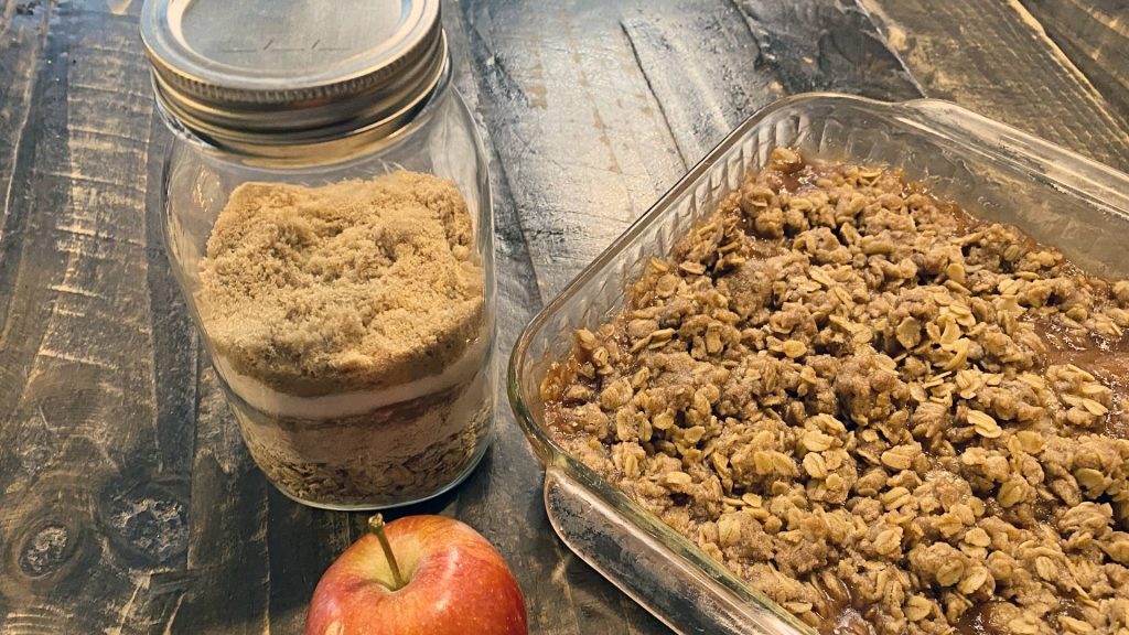 Apple crisp topping mix layered in a mason jar next to baked apple crisp in a glass dish on rustic wooden table