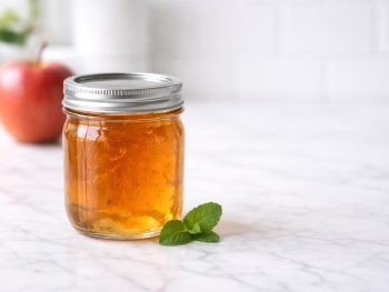 Translucent amber spiced apple mint jelly in a clear mason jar on white marble countertop with fresh mint and blurred red apple in background