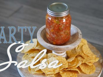 A glass jar filled with homemade party salsa sits on a wooden stand surrounded by crispy tortilla chips, ready for dipping.